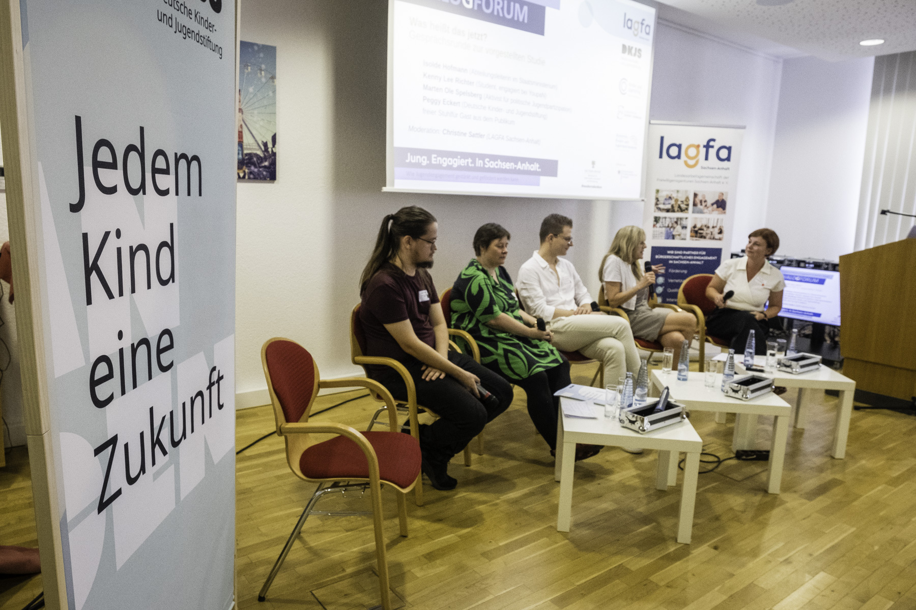 Fünf weiße Erwachsene, zwei Männer und drei Frauen, die in einem flachen Halbkreis vor einer Beamerleinwand sitzen, im Hintergrund das Logo der LAGFA.